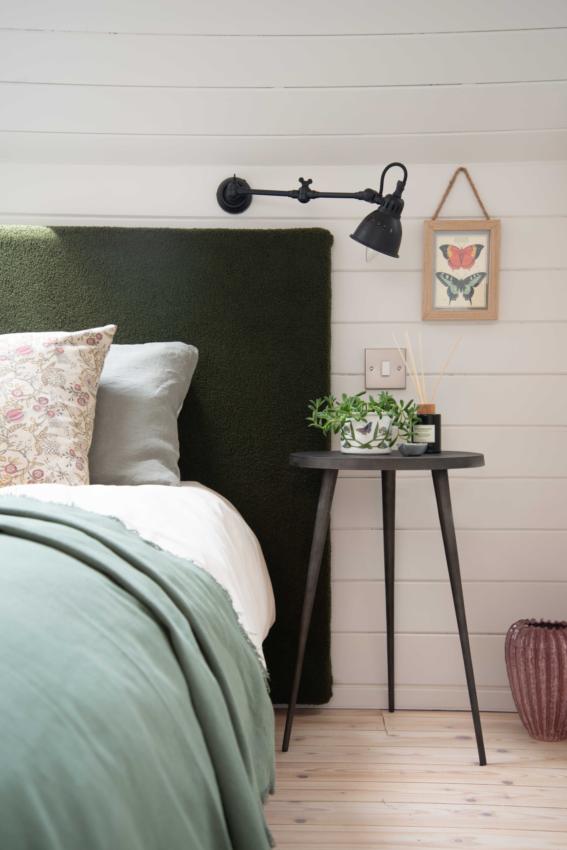 A custom made green headboard in boucle fabric, with rustic iron wall light and metal tripod style bedside table.