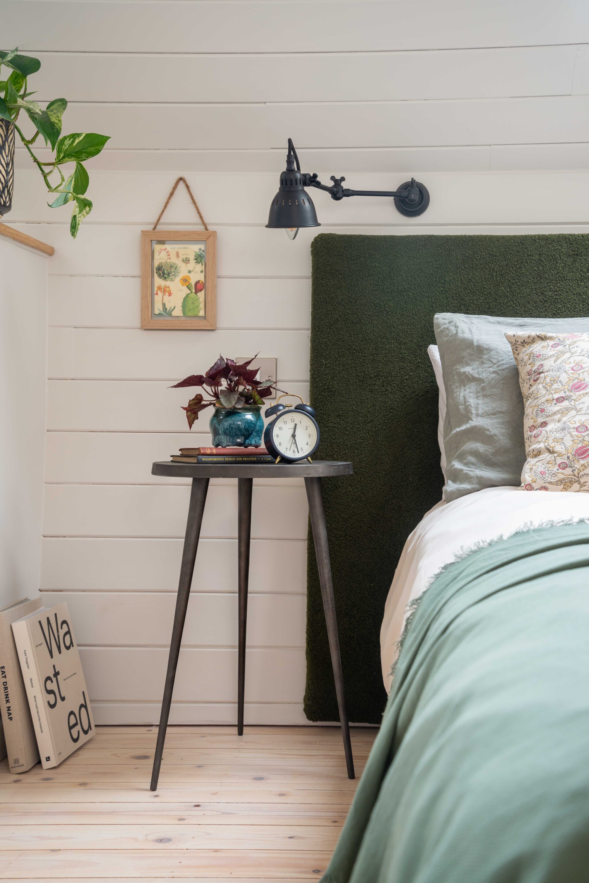 A custom made green headboard in boucle fabric, with rustic iron wall light and metal tripod style bedside table.