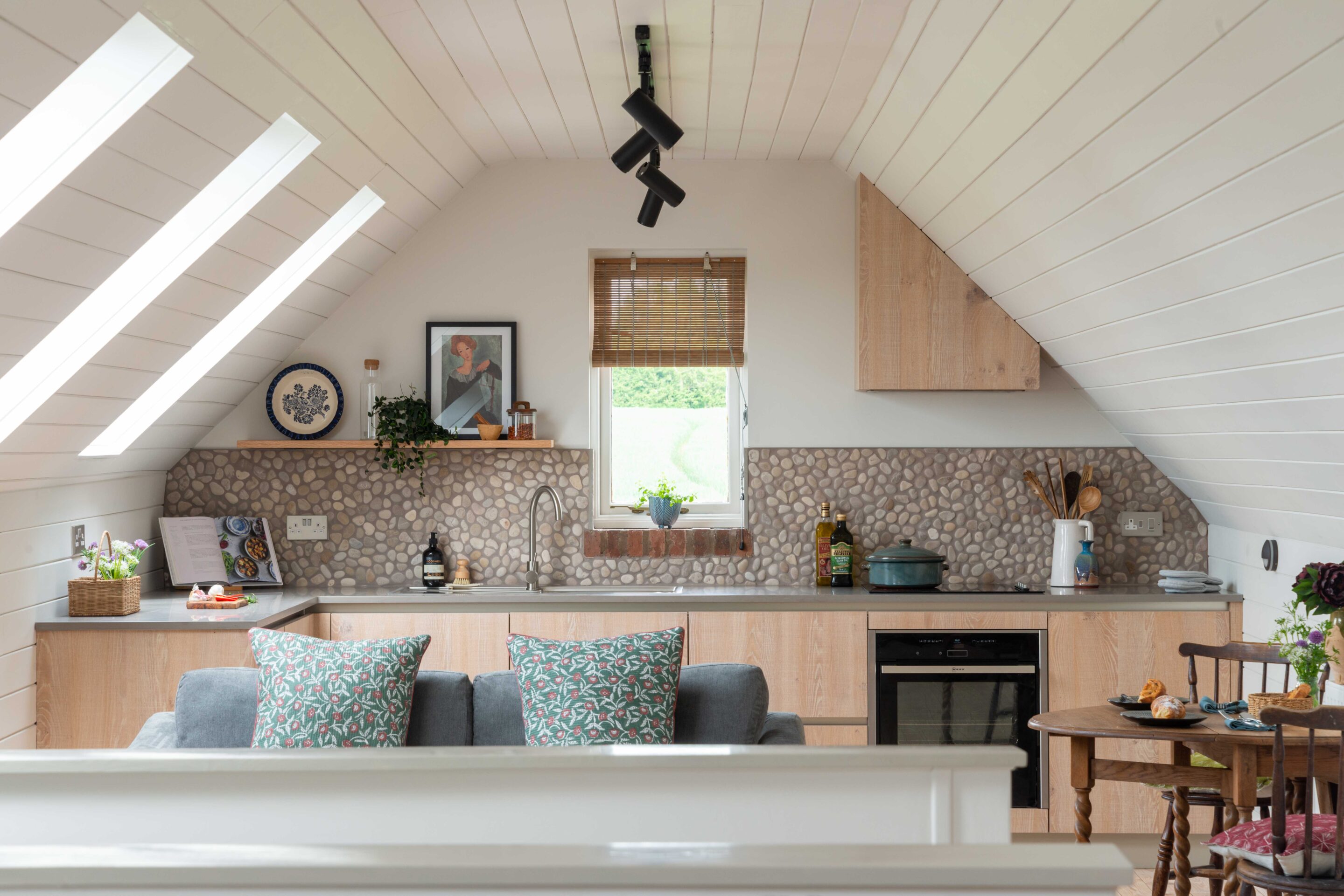 Images shows a bespoke kitchen and living space built into a loft conversation with oak veneer handless kitchen cabinets, a warm grey meganite worksuface and a quirky pebble backsplash.