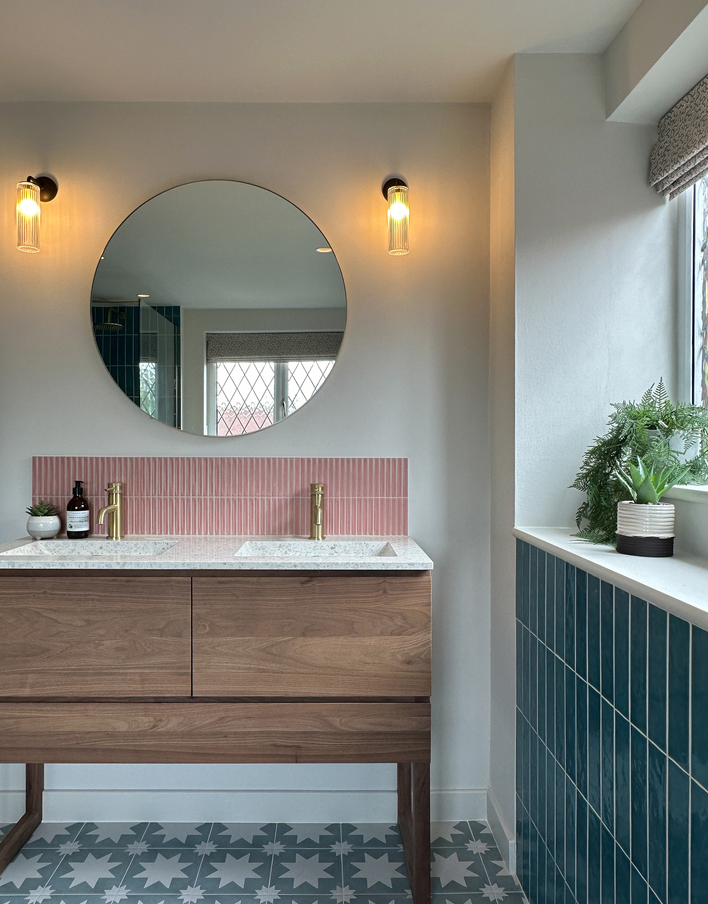 This picture shows a bathroom vanity unit in walnut with a terrazzo top. Pink tiles provide a splashback. A large round brass edged mirror sits above with fluted glass bathroom wall lights either side.