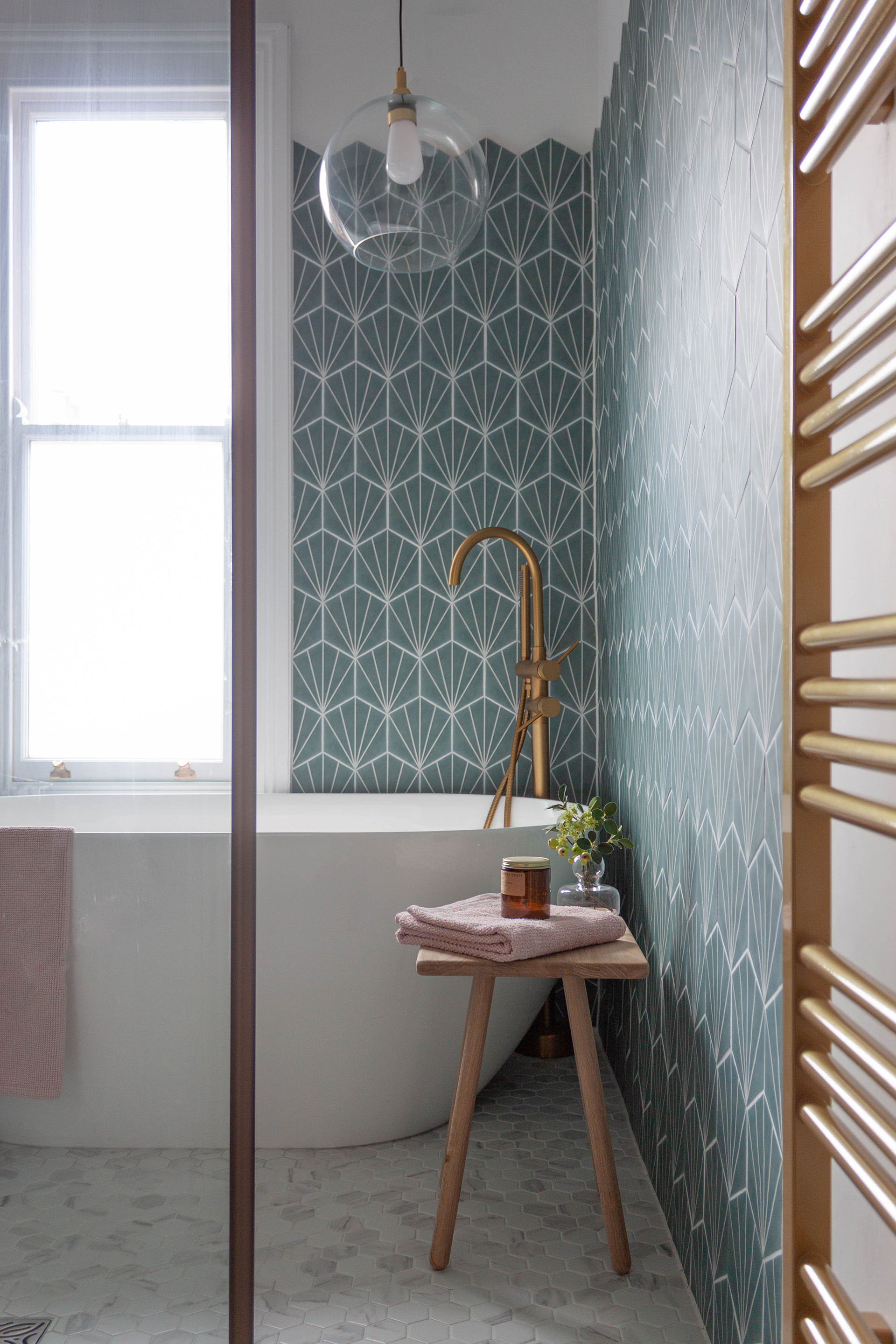 A snapshot of a wetroom area in a small victorian bathroom in Leamington Spa. Showing Hexagonal teal wall tiles, an elegant white freestanding bath and a brushed gold floorstanding bath filler. A bathroom rated pendant hangs over the bath.