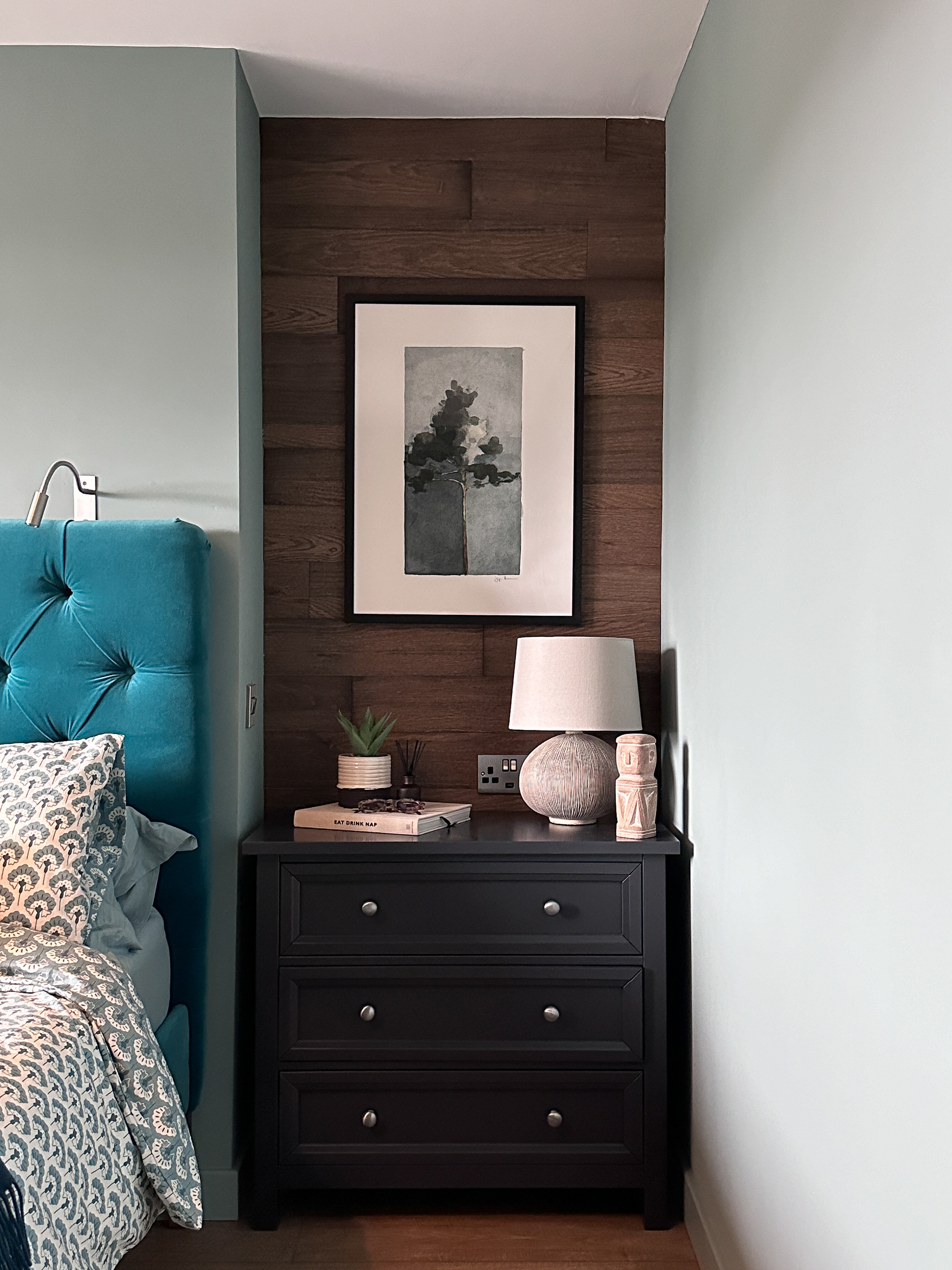 This picture shows a recess space beside the bed which has been panelled in rustic wood at the back, against walls painted in Farrow and Ball Oval Room Blue, a grey green blue, with a black chest of drawers and ceramic lamp.