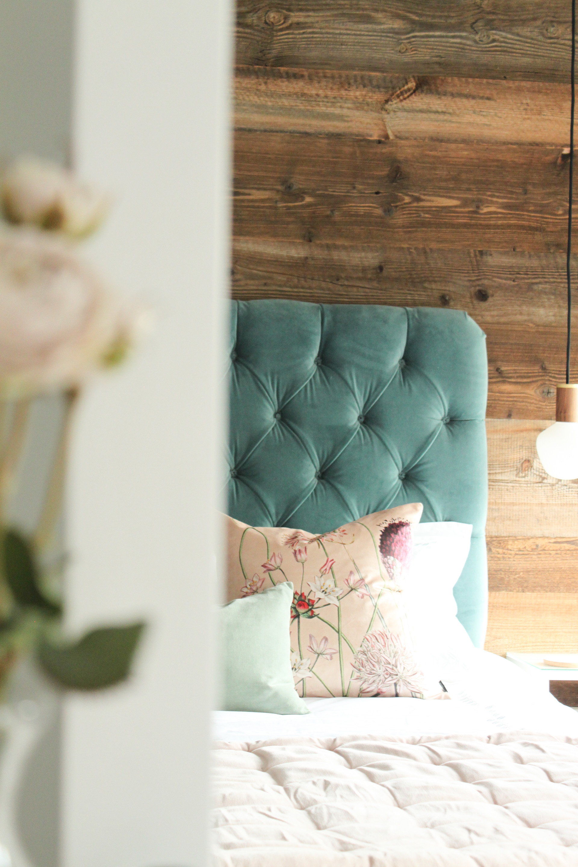 A reflected image of the rustic reclaimed wood floor to ceiling feature wall. A teal velvet headboard with bed dressing on a pale pink velvet throw and scatter cushions. A tala pendant light hangs from the ceiling above the bespoke bedside cabinets.