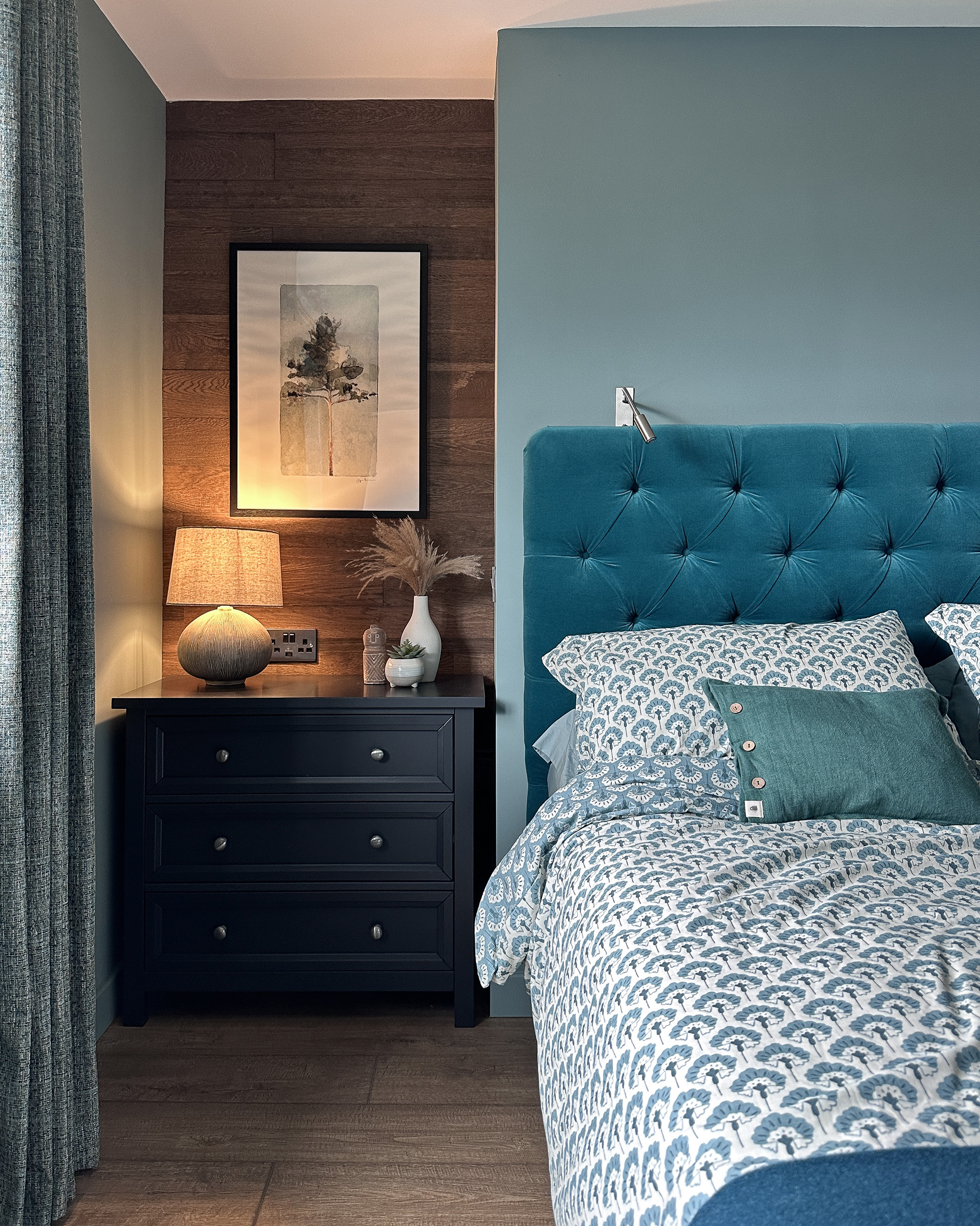 This picture shows a recess space beside the bed which has been panelled in rustic wood at the back, against walls painted in Farrow and Ball Oval Room Blue, a grey green blue, with a black chest of drawers and ceramic lamp. The bed has a teal velvet headboard and is dressed in a motif pattern duvet set from John Lewis.