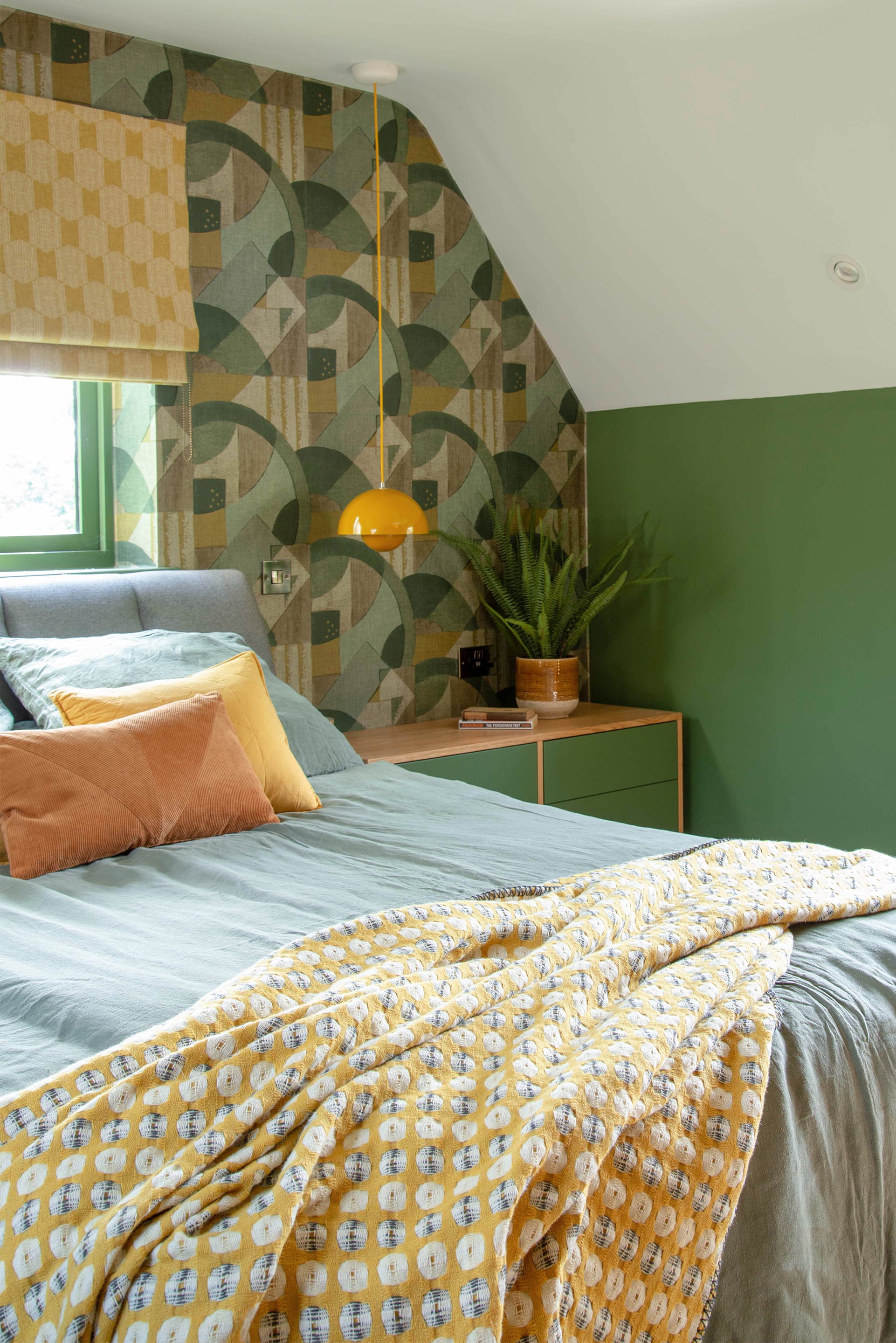 A colourful mid-century influenced bedroom, with abstract green and gold wallpaper, rich green walls and vibrant yellow pops of colour in the lamps and blankets. 