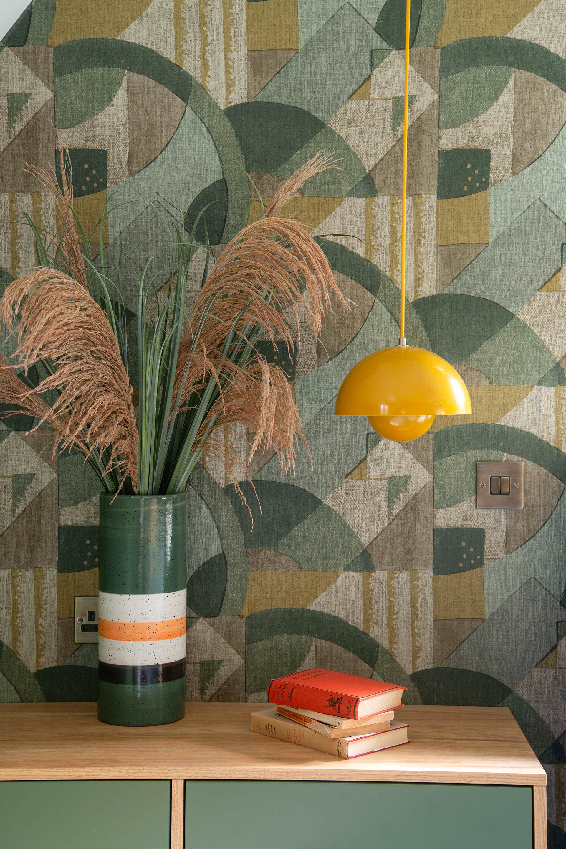Close up of a bedside table and abstract wallpaper in a mid-century style bedroom. Wallpaper is green and golds, with the bedside table in oak veneer and green drawer fronts. A bright yellow &tradition Flower Pot Pendant hangs above. 