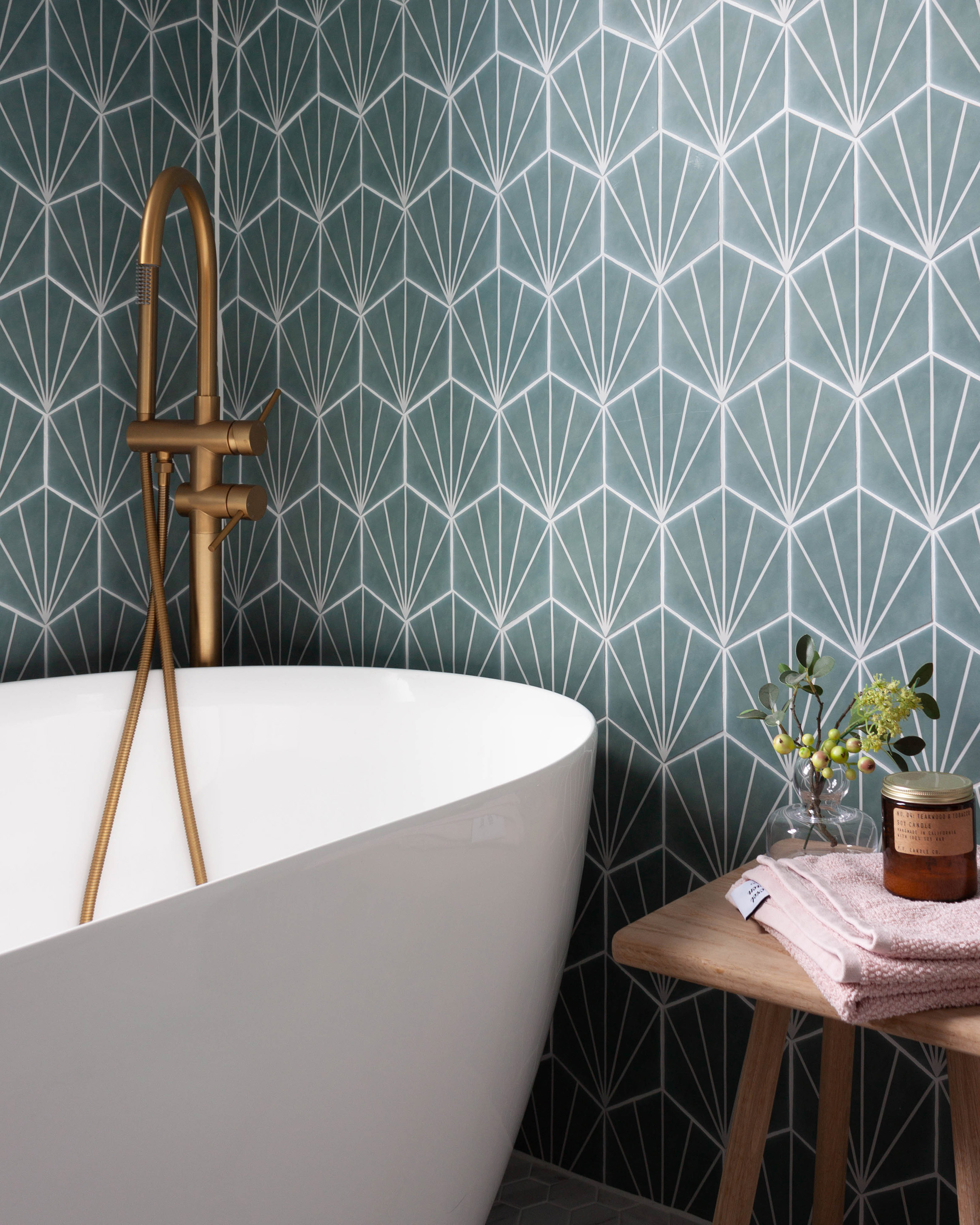 An elegant white freestanding bath with a gold floor mounted bath filler and shower head, set against vibrant teal hexagonal tiles with a white starburst pattern. A section of a bathroom design in Leamington Spa.