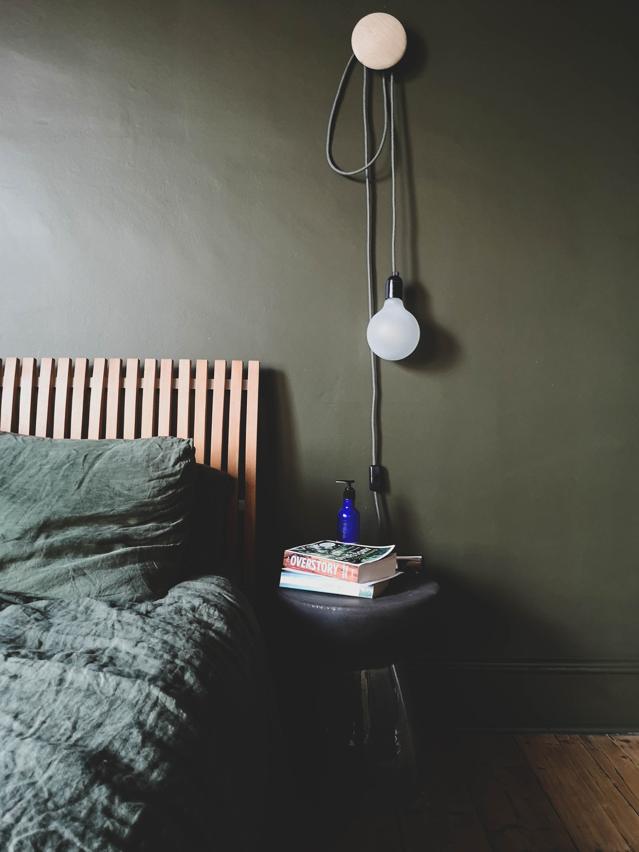 A picture of a bed against the dark green walls of the re-designed bedroom. The bumpy rough texture of the original walls in this victorian terrace add to the rustic, crumpled character of the room. A simply decorative bulb hangs from a hook beside the bed over a ceramic bedside table.
