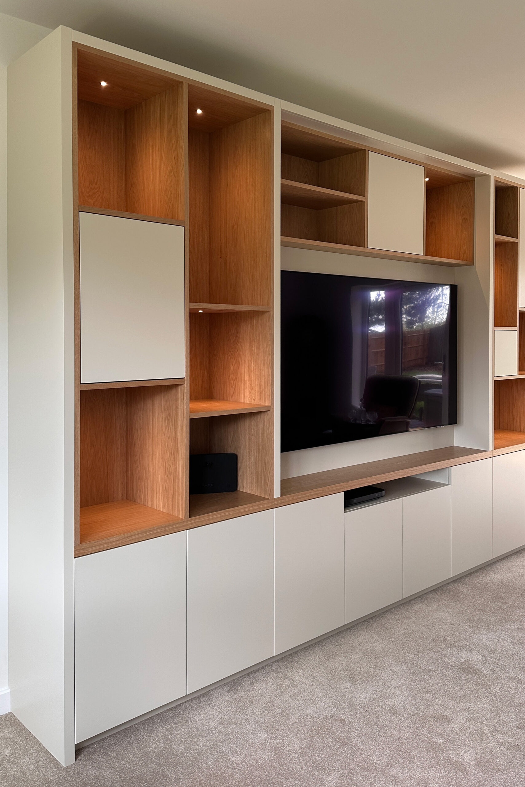 Projects 2 A large bespoke media wall made from Oak Veneer and Spray Painted in Little Greene Portland Stone. The cabinetry houses a large TV with a mixture of open and closed shelving and storage in an asymetric design. TV Cables are all hidden.