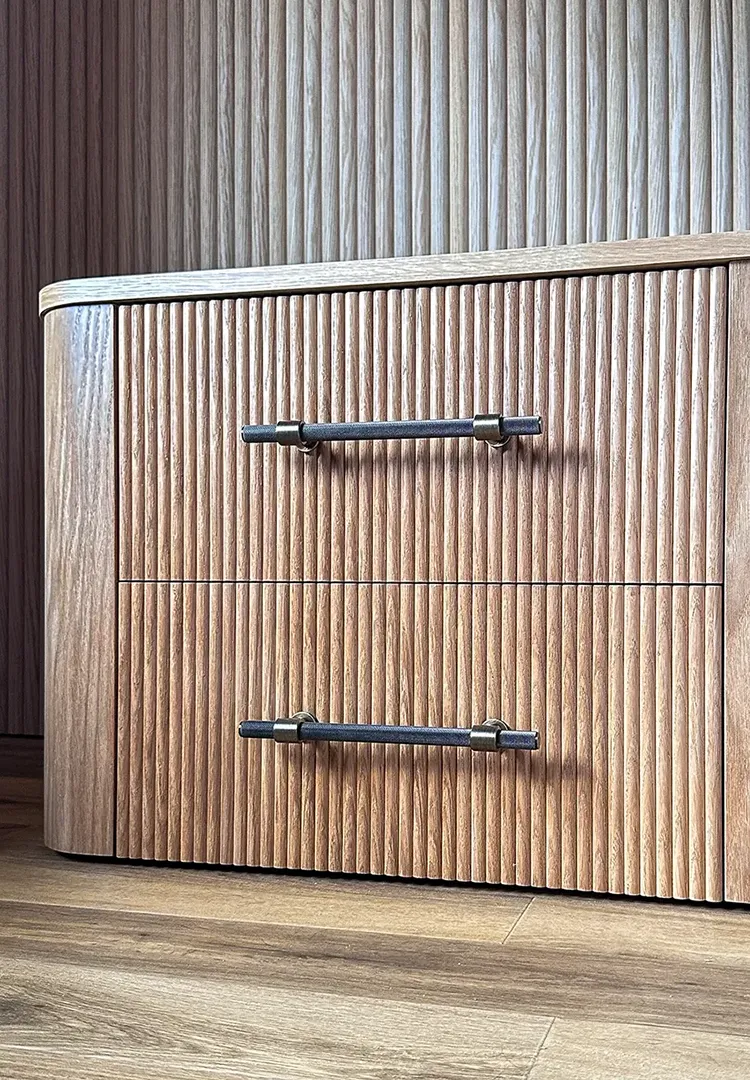 Close-up of a handcrafted oak bedside cabinet with continuous vertical reeding, rounded corners and dark brass drawer handles.