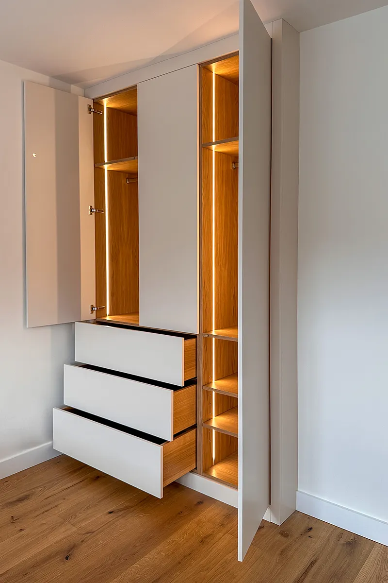 Open bespoke fitted wardrobe with oak interiors, integrated warm lighting, hanging space, shelving and internal drawers.