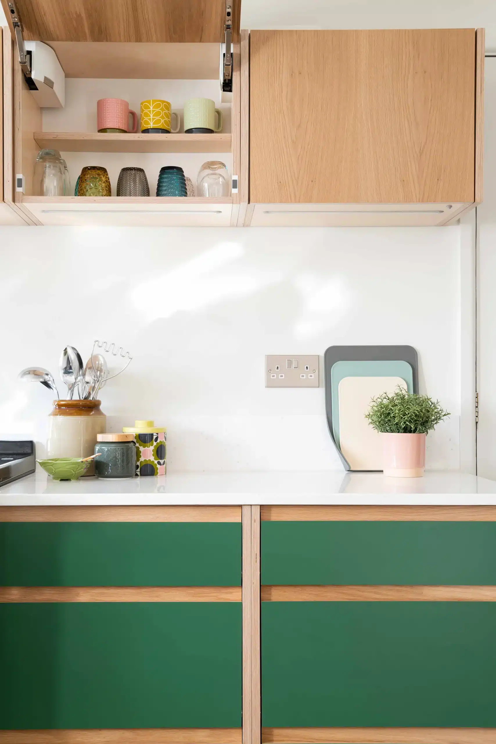 Bespoke green kitchen base cabinets with natural oak detailing and lift-up wall cupboards displaying colourful mugs and glasses.