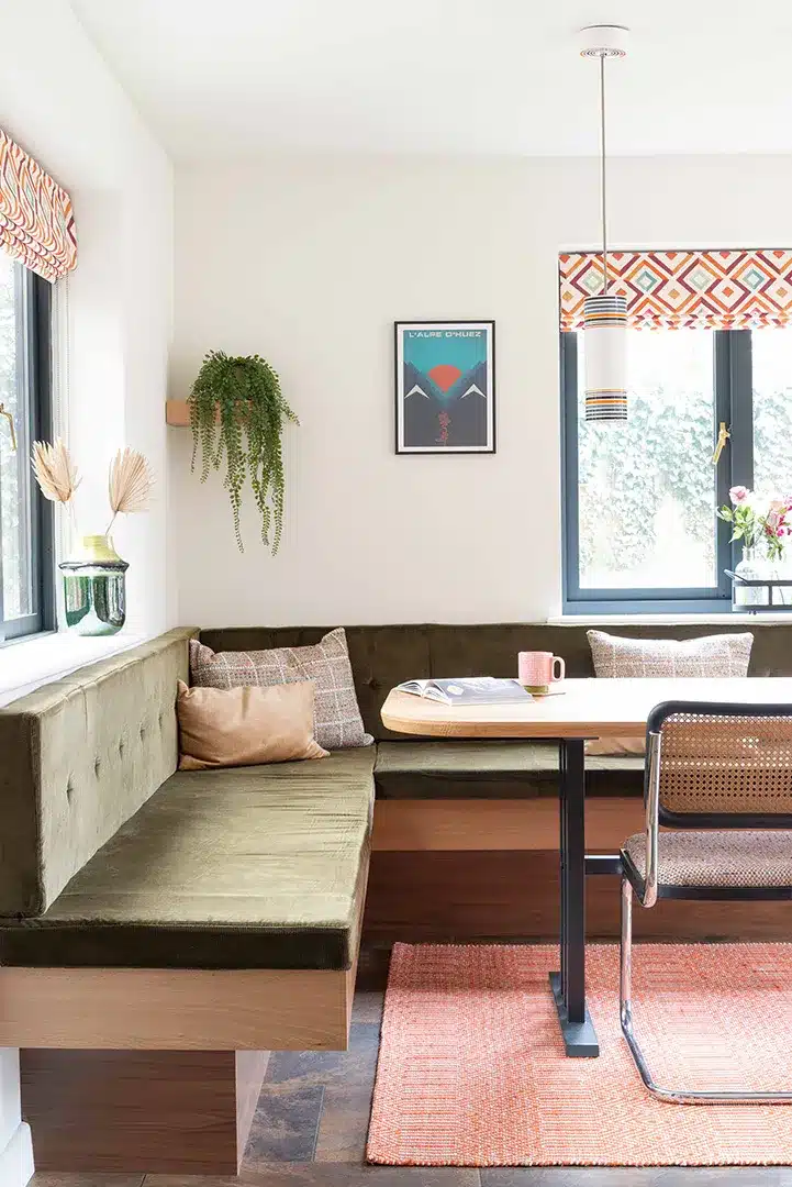 Bespoke L-shaped dining banquette with olive green upholstered seating, timber base and dining table beside two windows.
