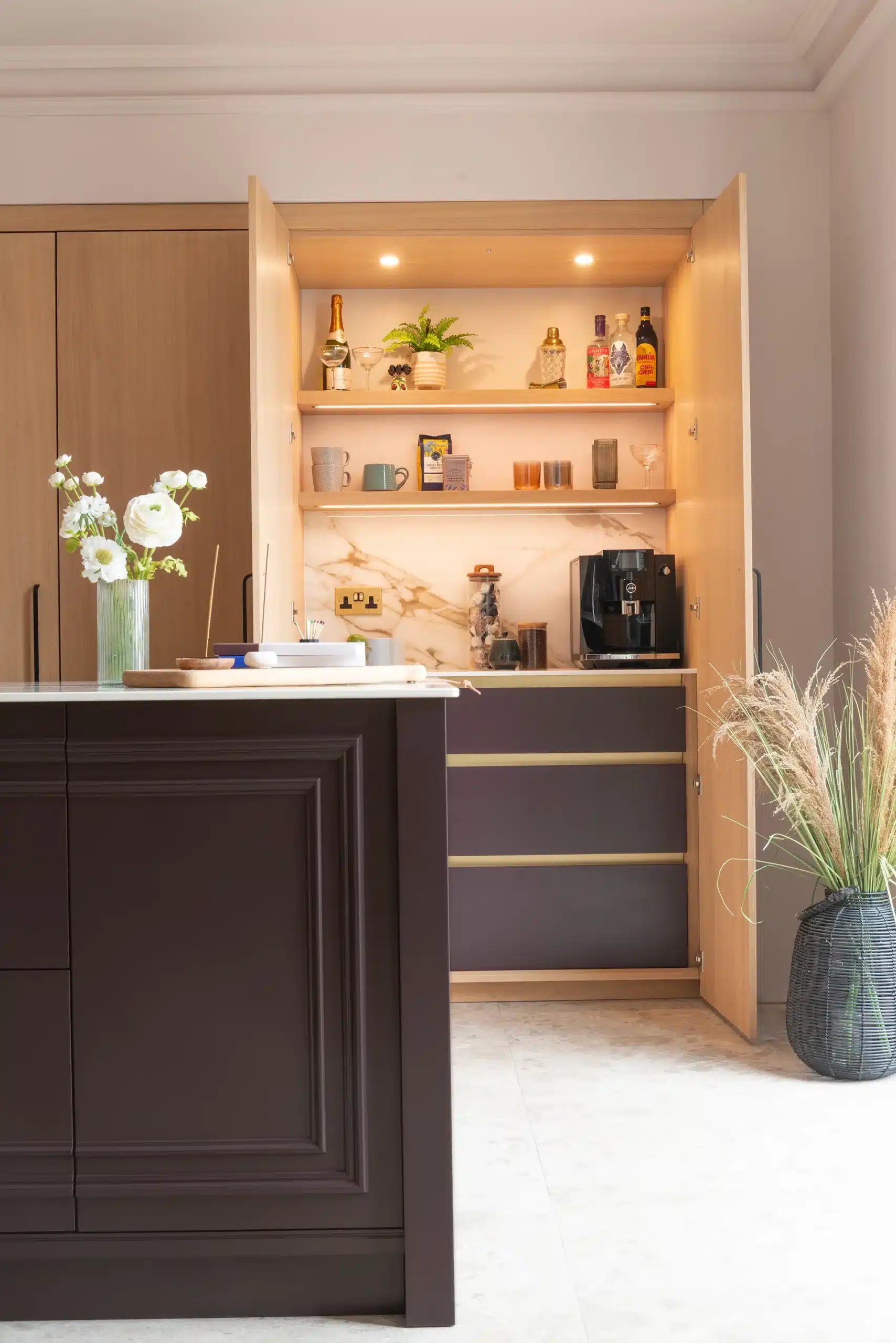 Open oak breakfast cupboard with integrated lighting, shelves, coffee machine and internal drawers beside a dark painted kitchen island.
