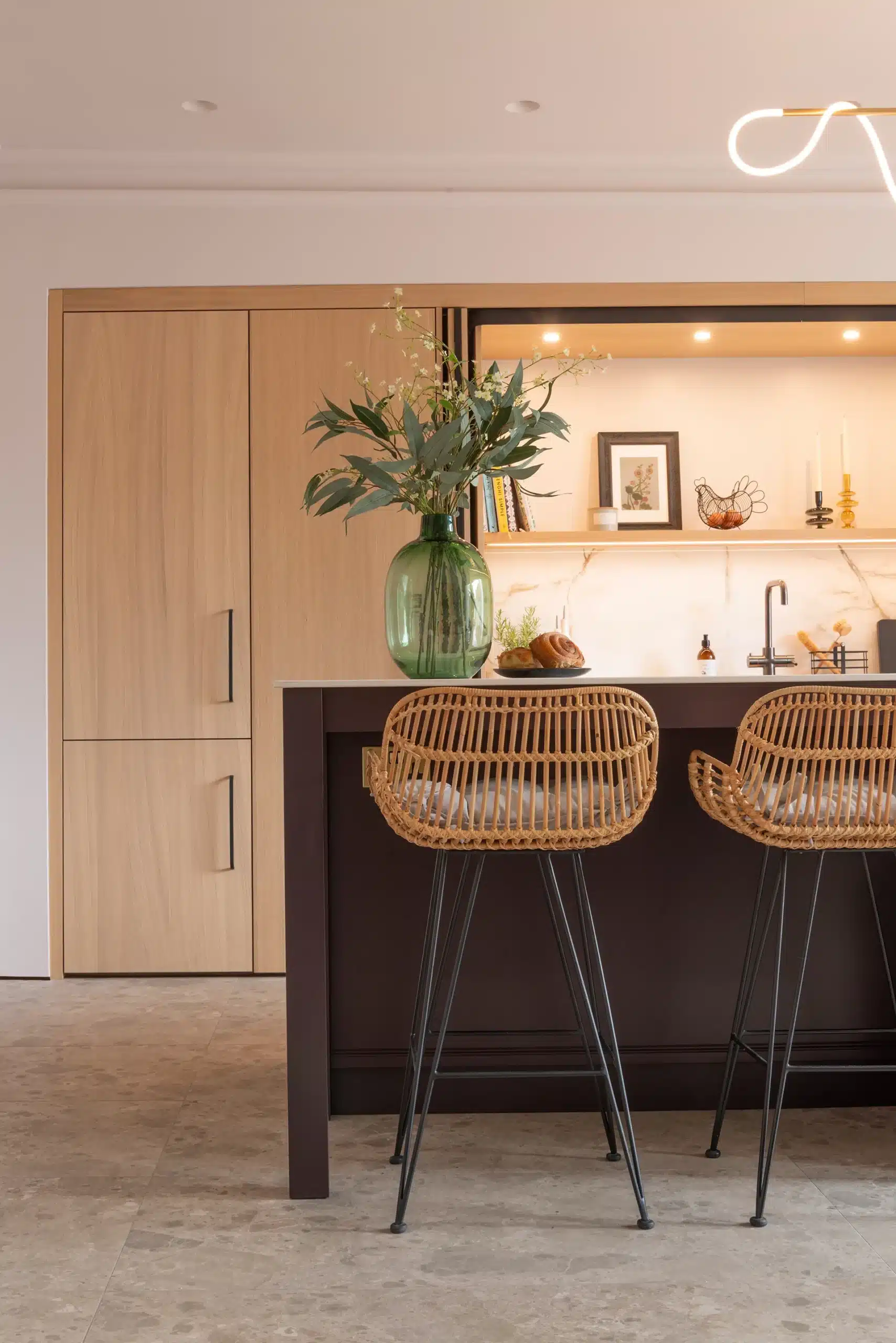 Dark painted kitchen island with woven breakfast bar stools, full-height oak cabinetry and illuminated shelving behind.